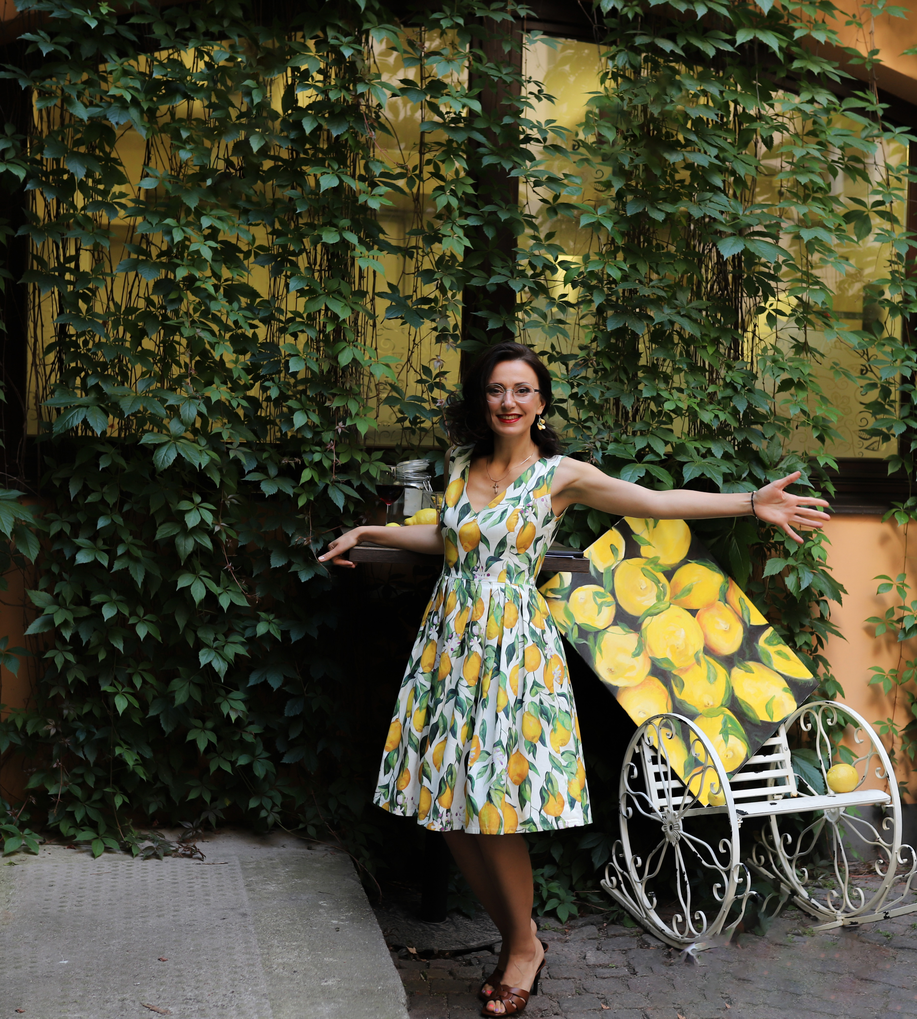 Sieviete košā kleitā ar dzelteniem citroniem stāv pie zaļu augu sienas