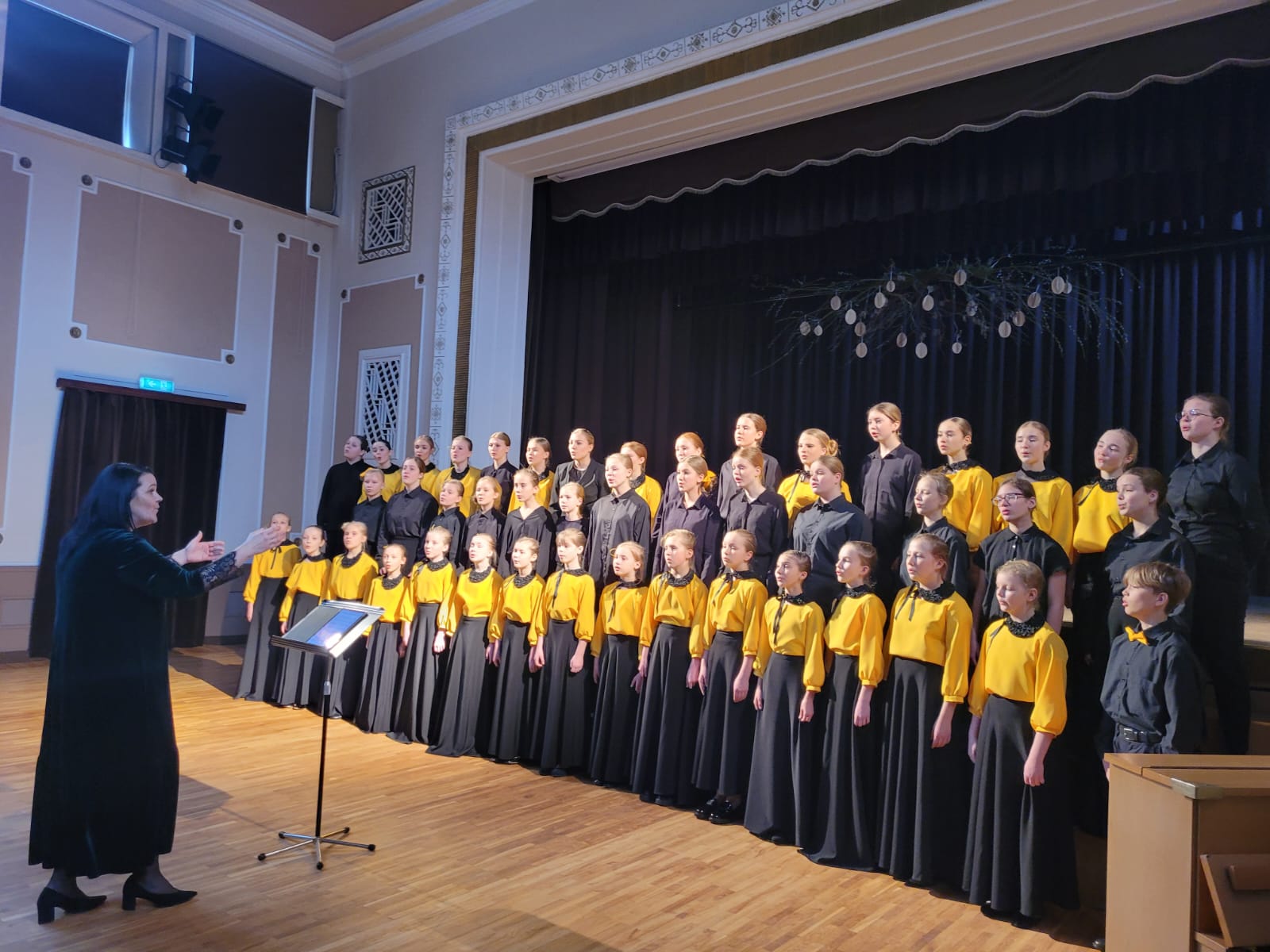 Skolēnu koris dzelteni melnos tērpos stāv uz skatuves un diriģē vadītāja