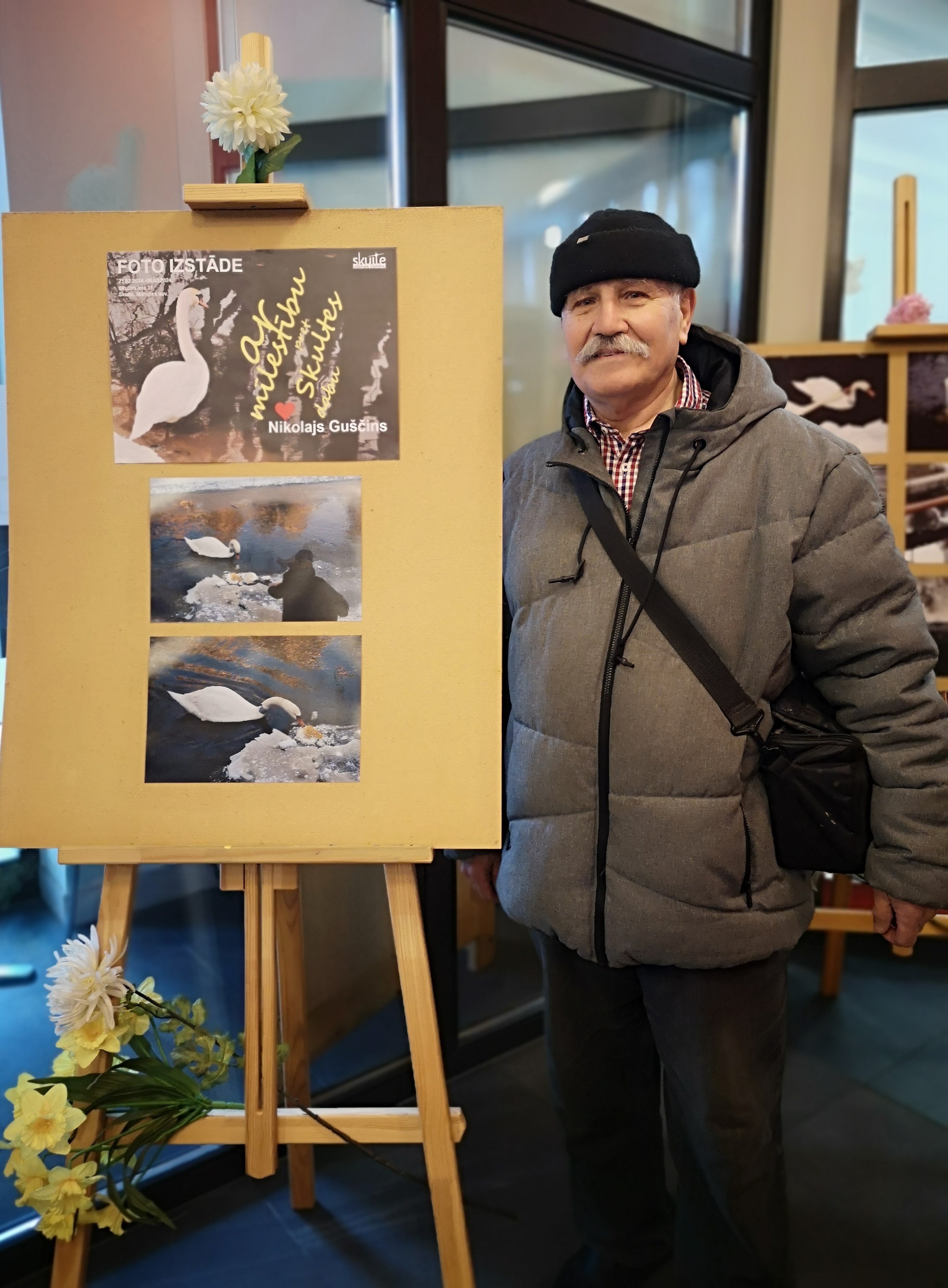 Skultenieks Nikolajs Guščins pie molberta ar plakātiem par savu foto izstādi dienas centrā Skultē