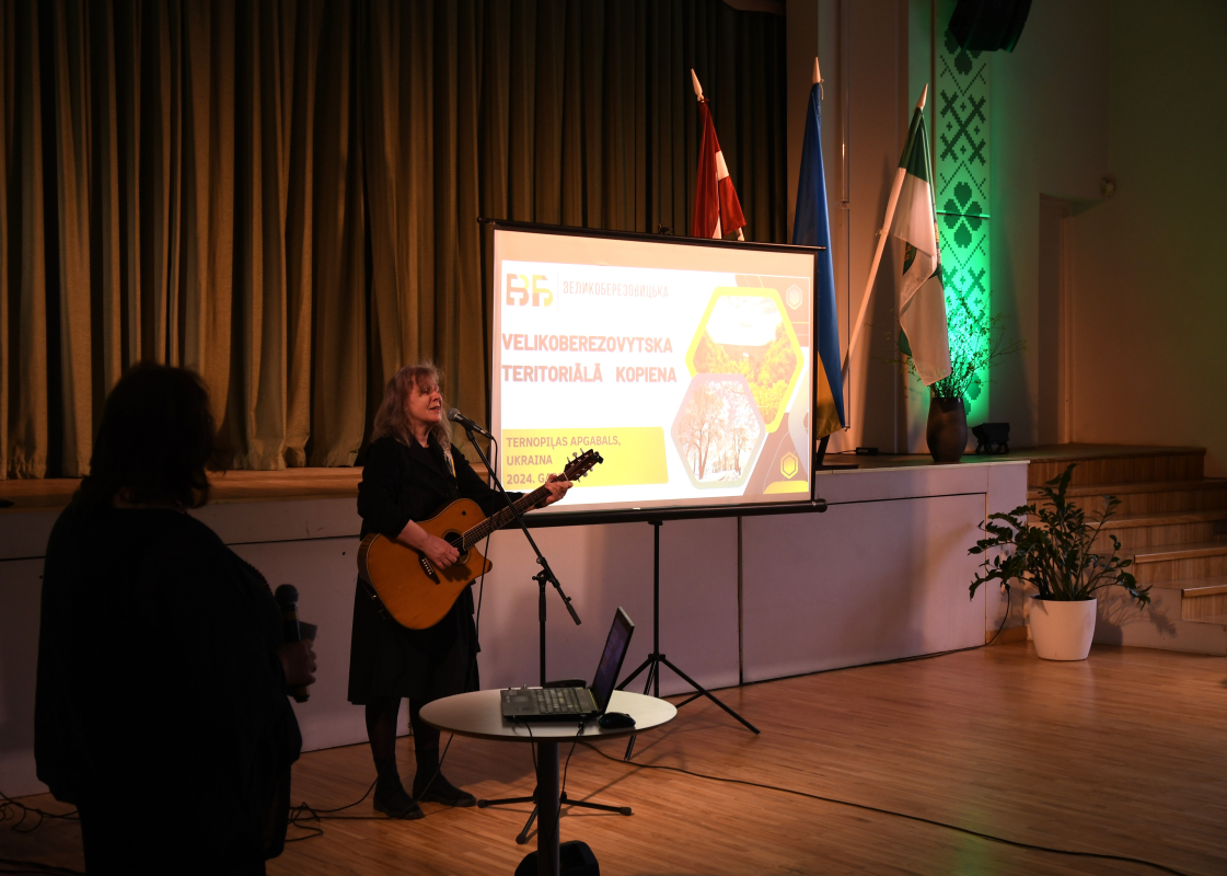 Sieviete ar ģitāru rokās dzied uz skatuves, blakus projektora ekrāns
