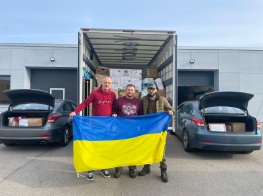 Trīs vīrieši stāv pie kravas busa, ar Ukrainas karogu rokās