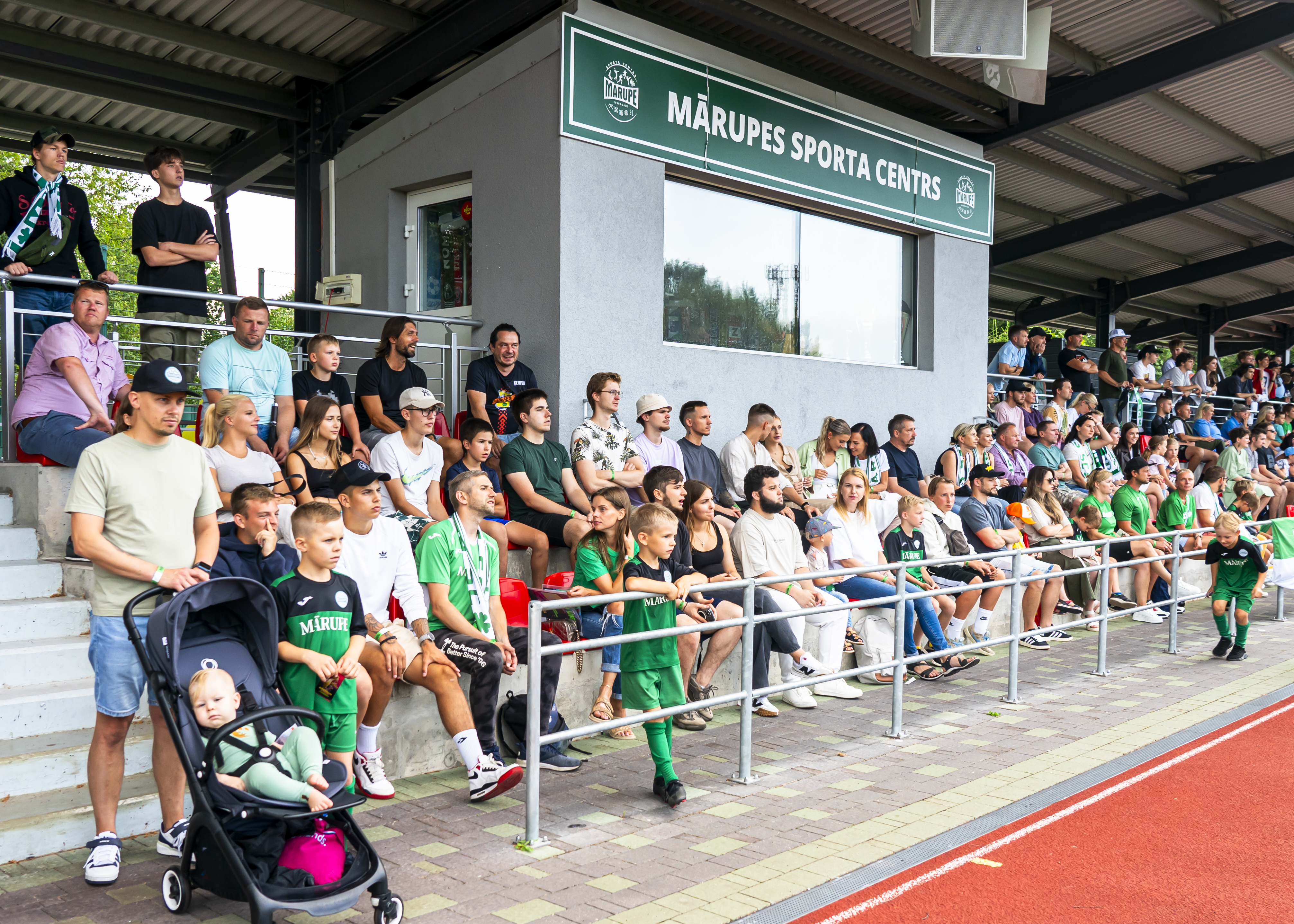 Daudz cilvēku sēž tribīnēs, futbolistu līdzjutēji