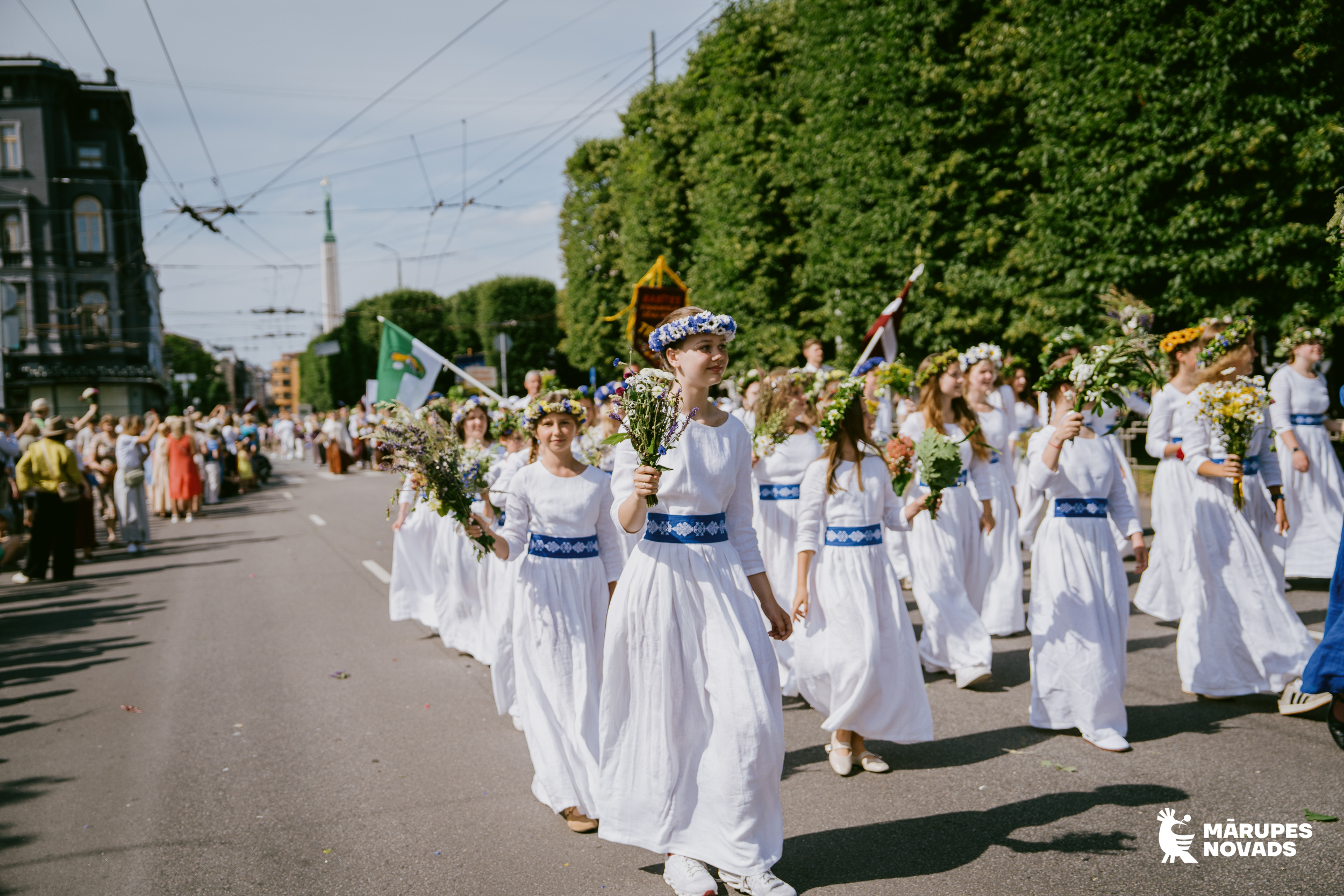 Meitenes baltās kleitās un vainagiem galvā piedalās gājienā