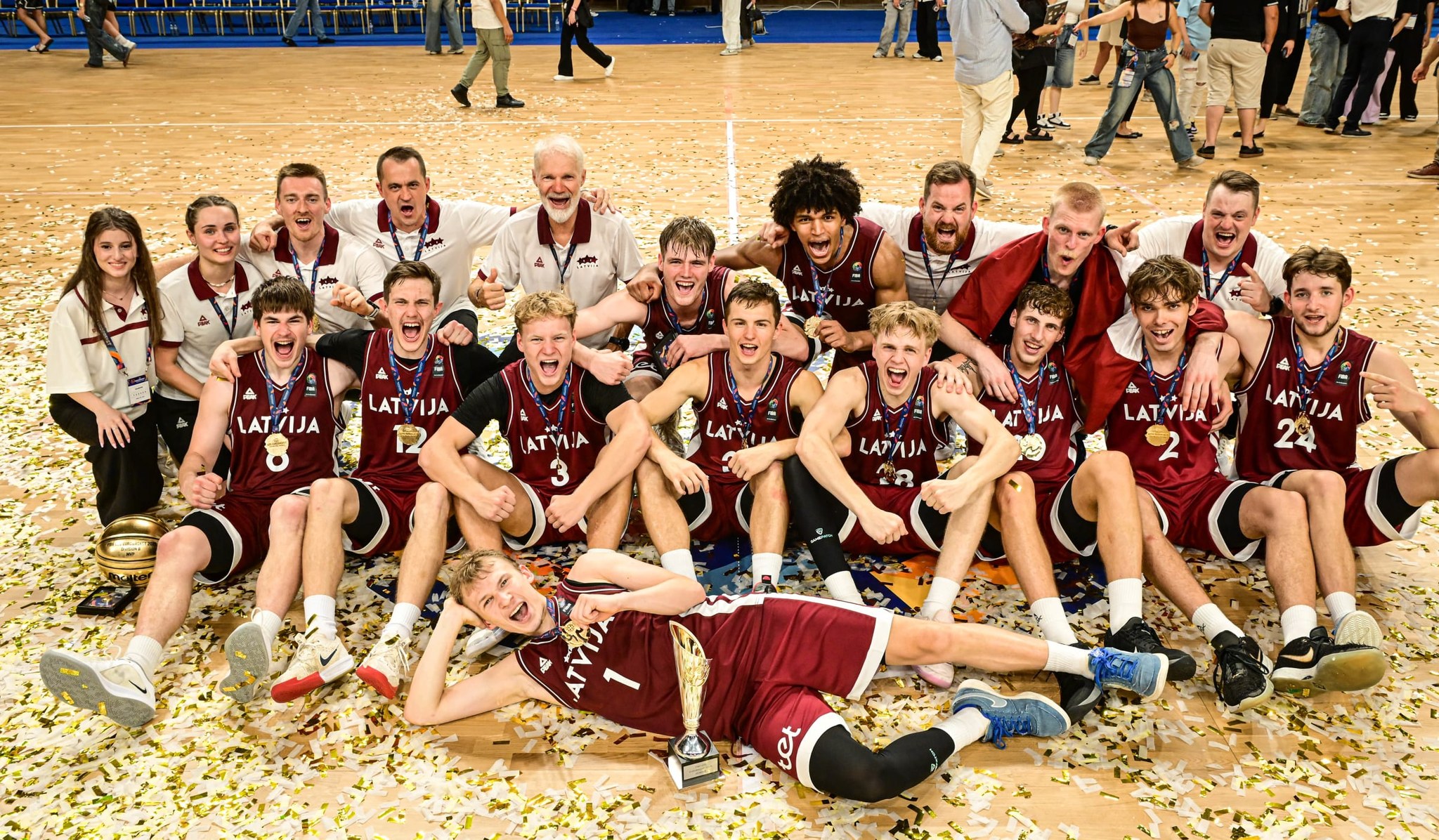 Basketbolisti sarkanos tērpos uz zāles ar kausu priekšā
