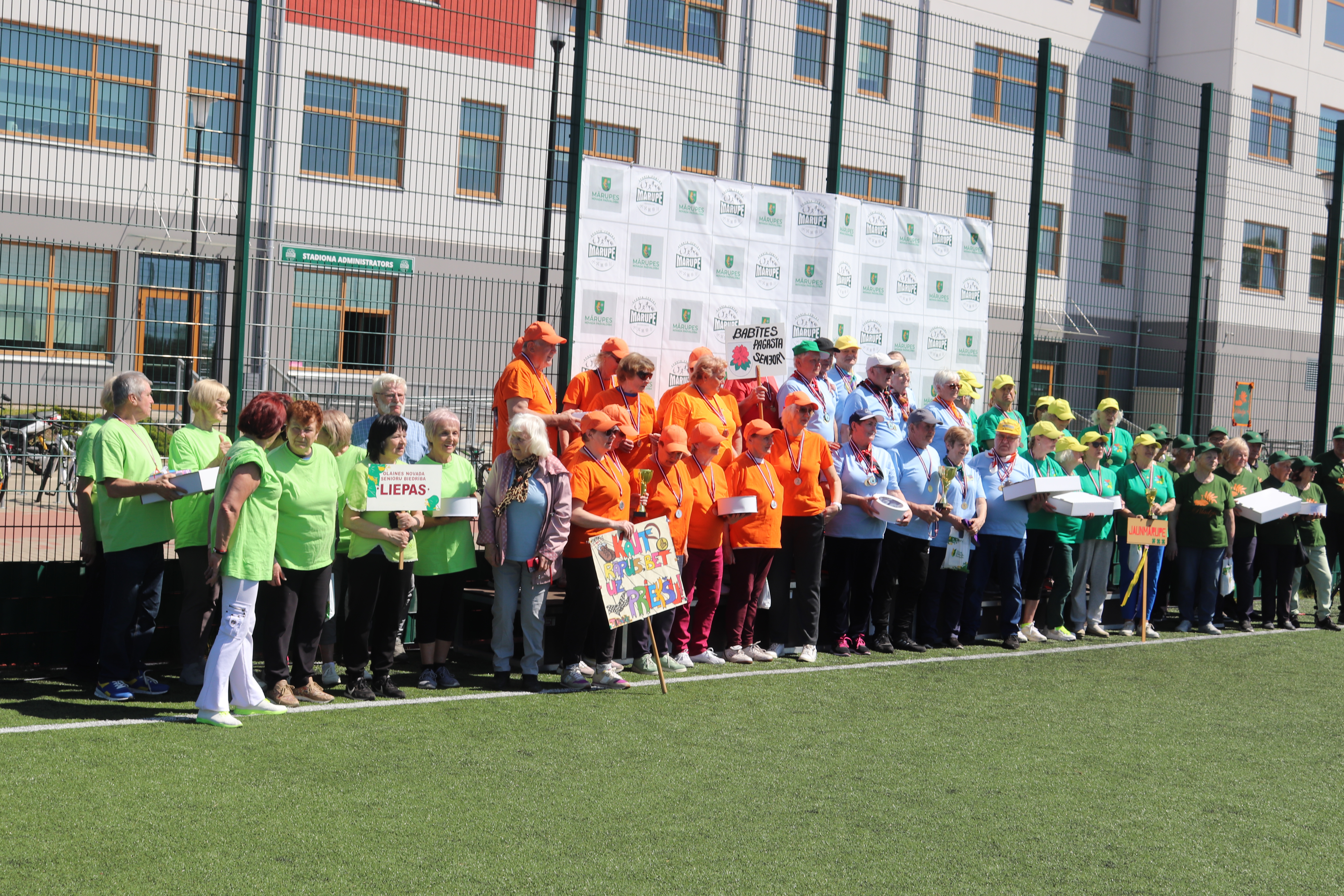 seniori stāv pie foto sienas futbola laukumā
