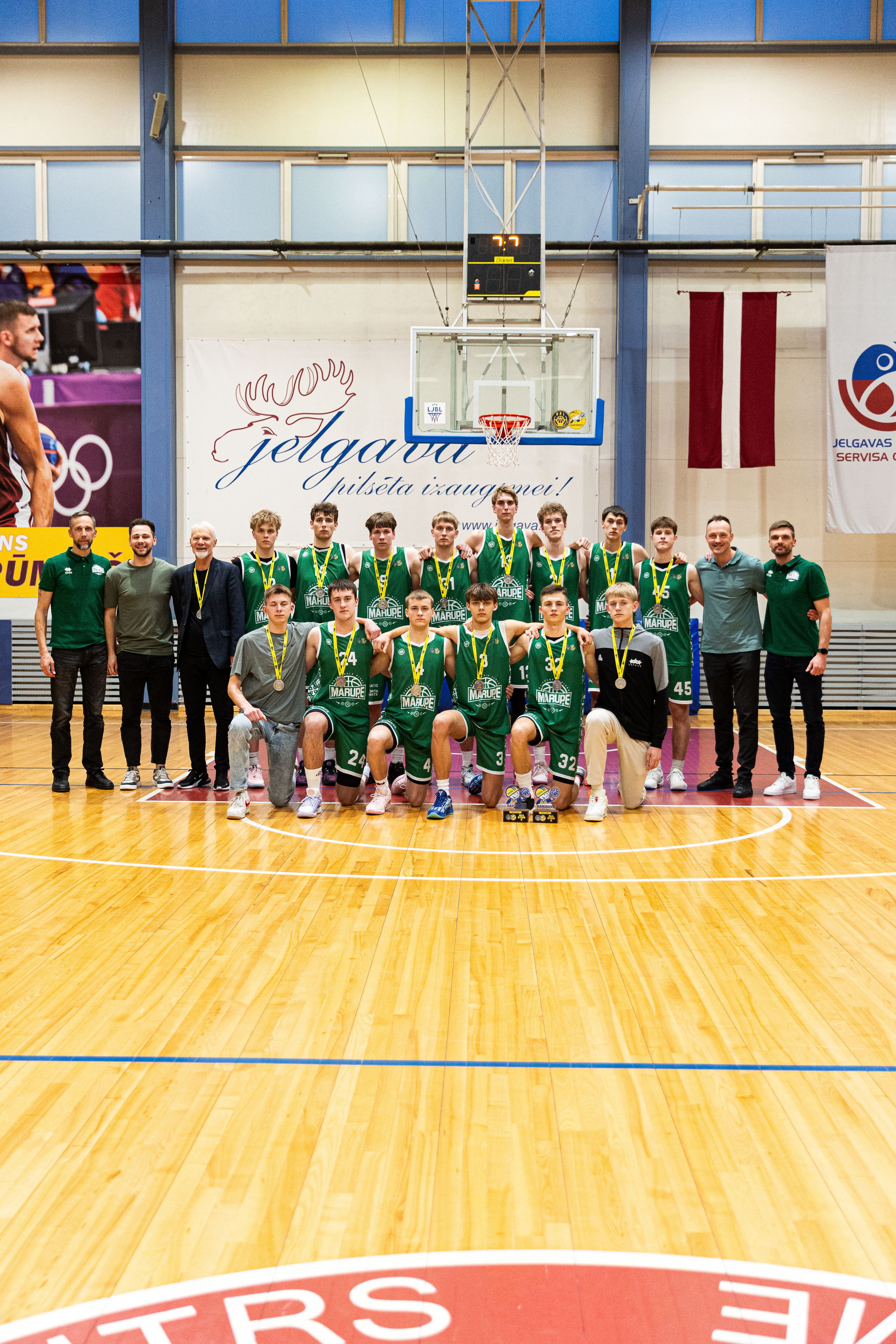 Vairāki puiši un vīrieši stāv sporta zālē ar sudraba medaļām kaklā