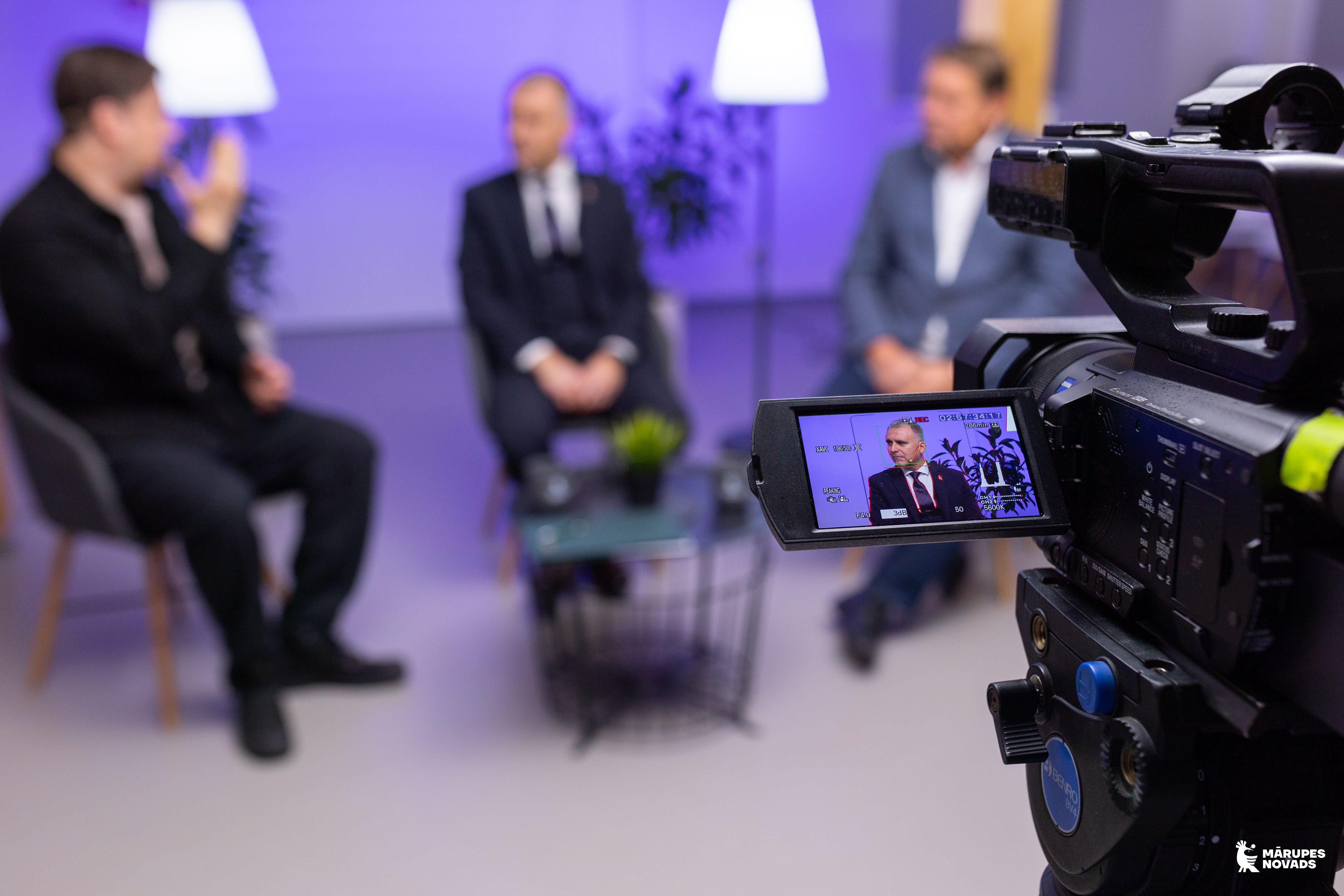 Video kameras ekrāna tuvplāns, kurā redzams vīrietis melnā uzvalkā