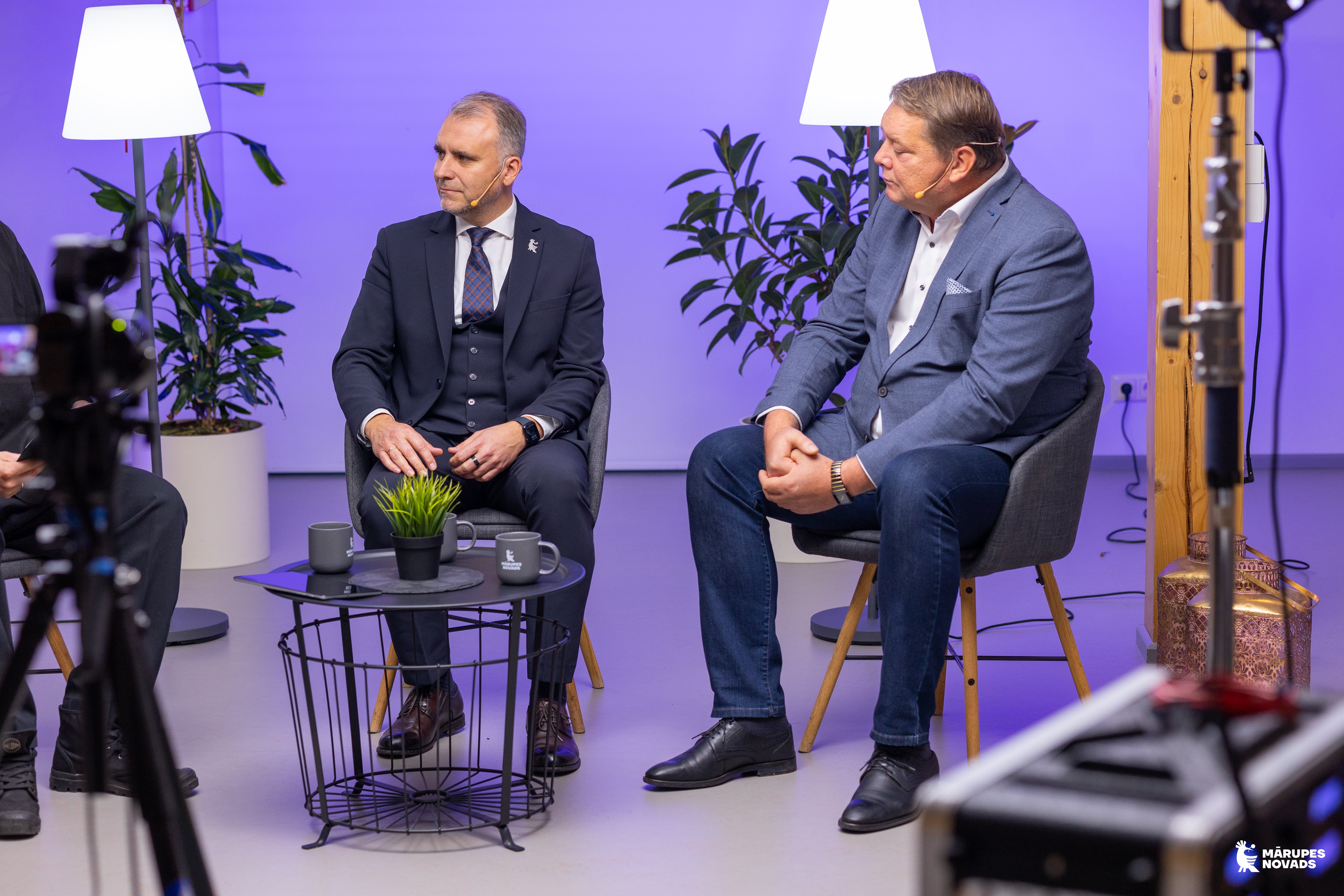 Divi vīrieši uzvalkos sēž pelēkos krēslos