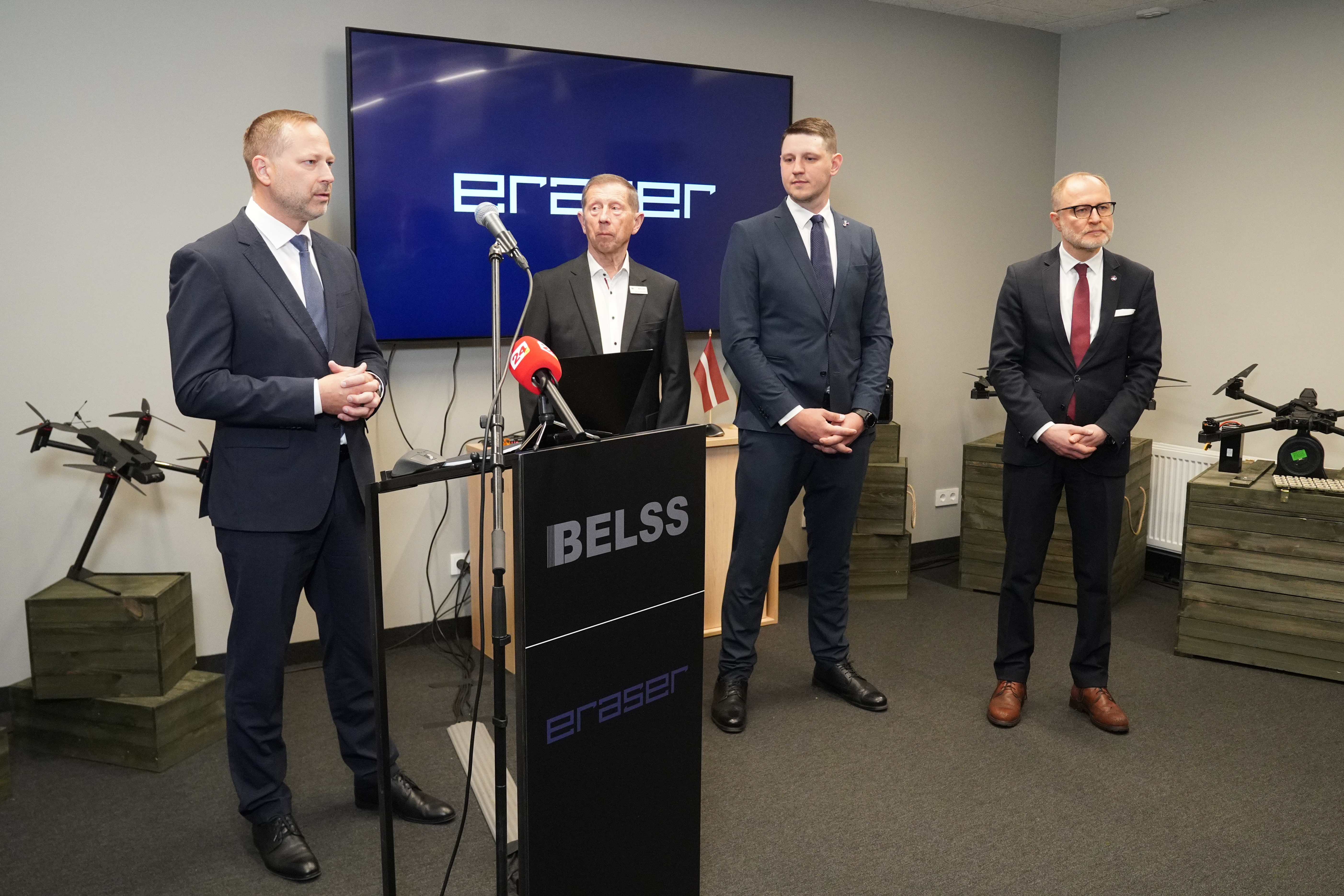Vīrieši uzvalkos preses konferencē pie mikrofoniem