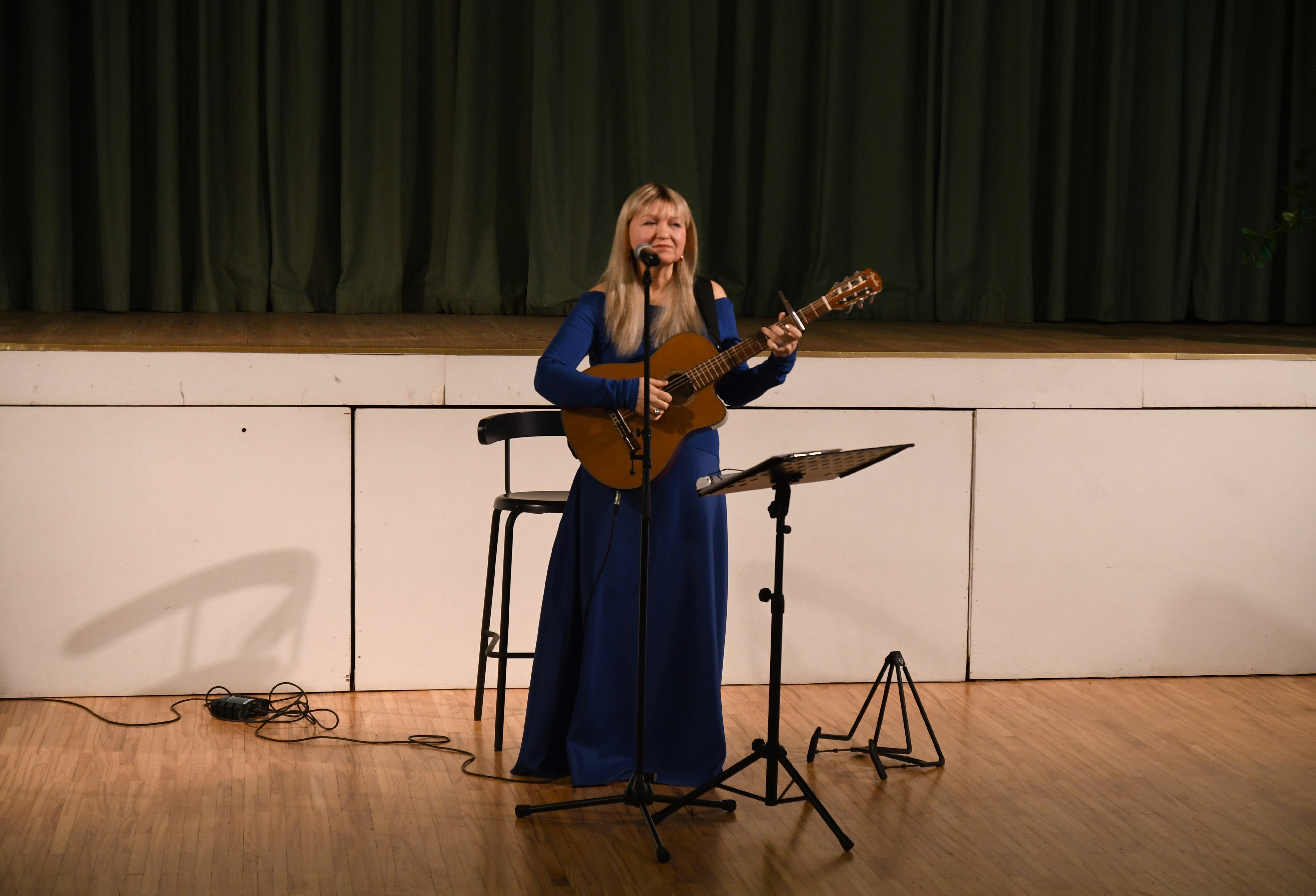 Sieviete zilā garā kleitā ar ģitāru pie mikrofona stāv un dzied