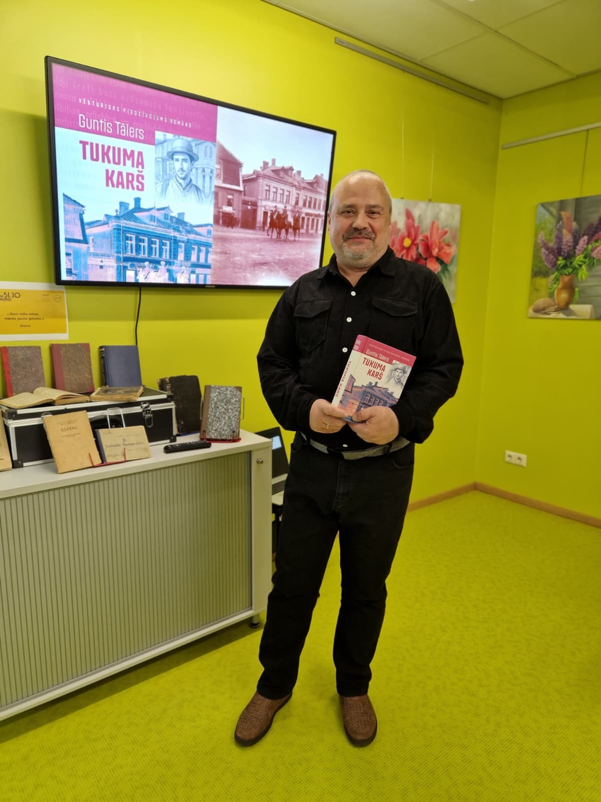 V\irietis tumšā uzvalkā ar grāmatu rokās telpā ar koši dzeltenām sienām