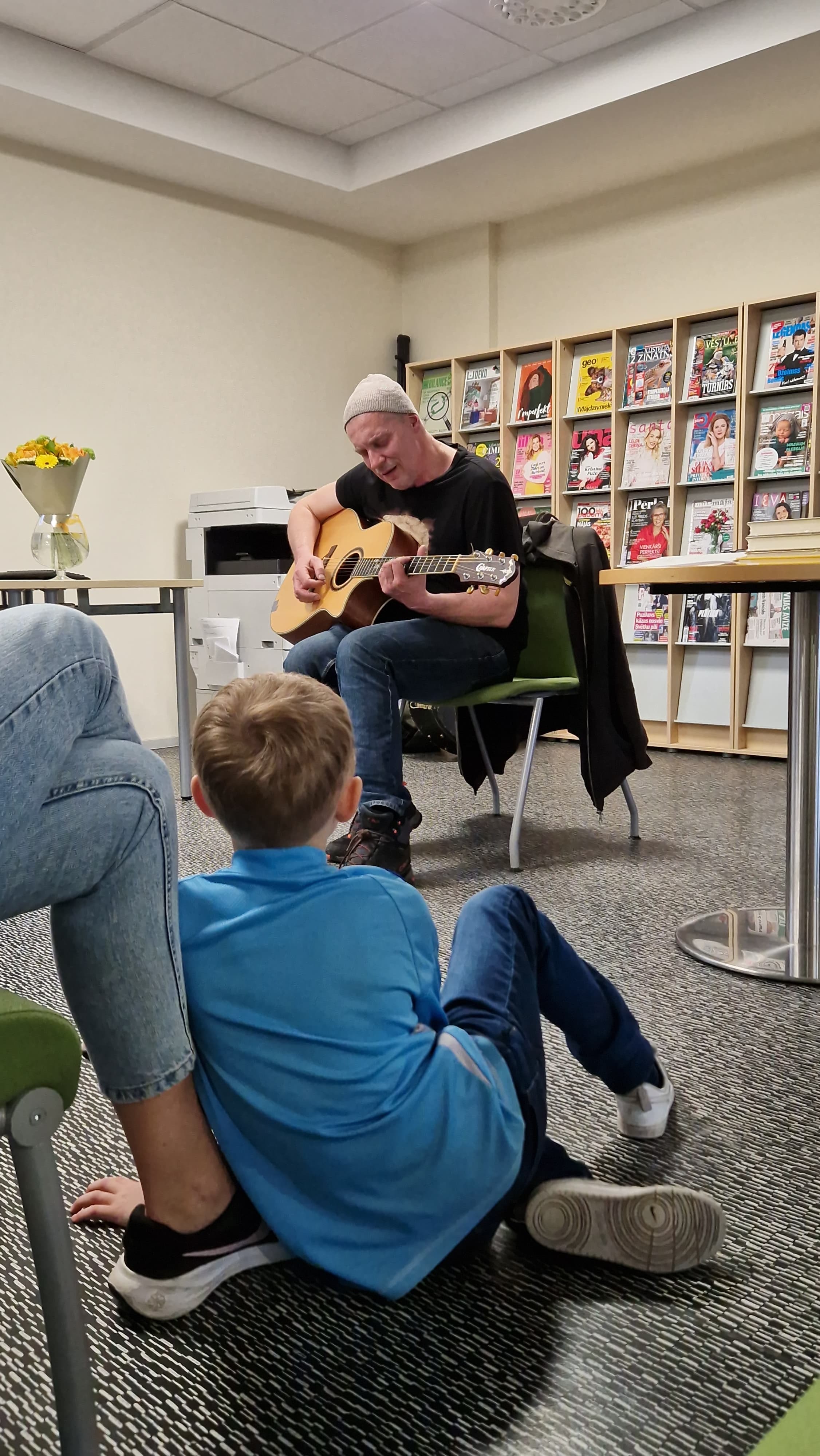 Vīrietis ar ģitāru muzicē apmeklētājiem gaišā telpā