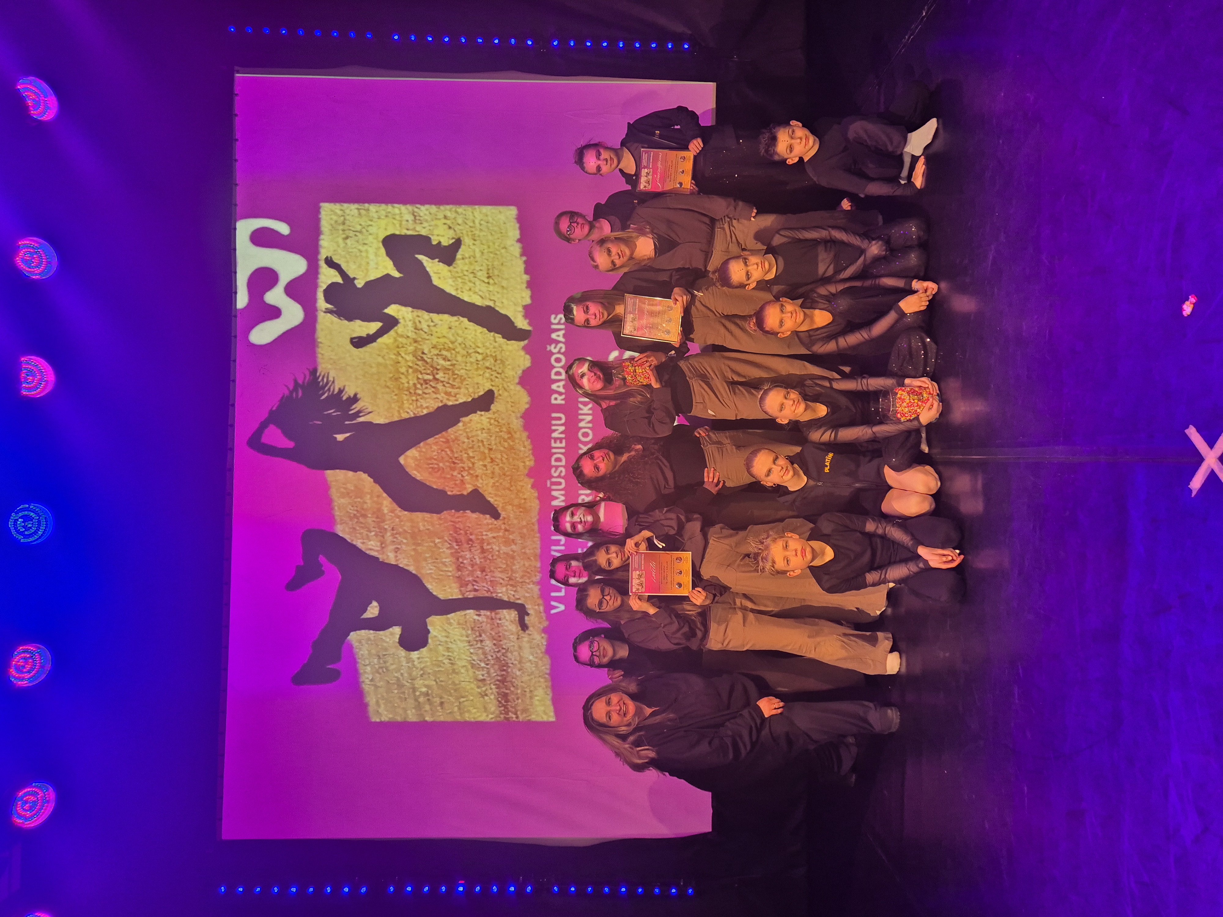 violetas noskaņas bilde, kur redzami bērni un jaunieši uz skatuves, kurus apspīd violeta gaisma