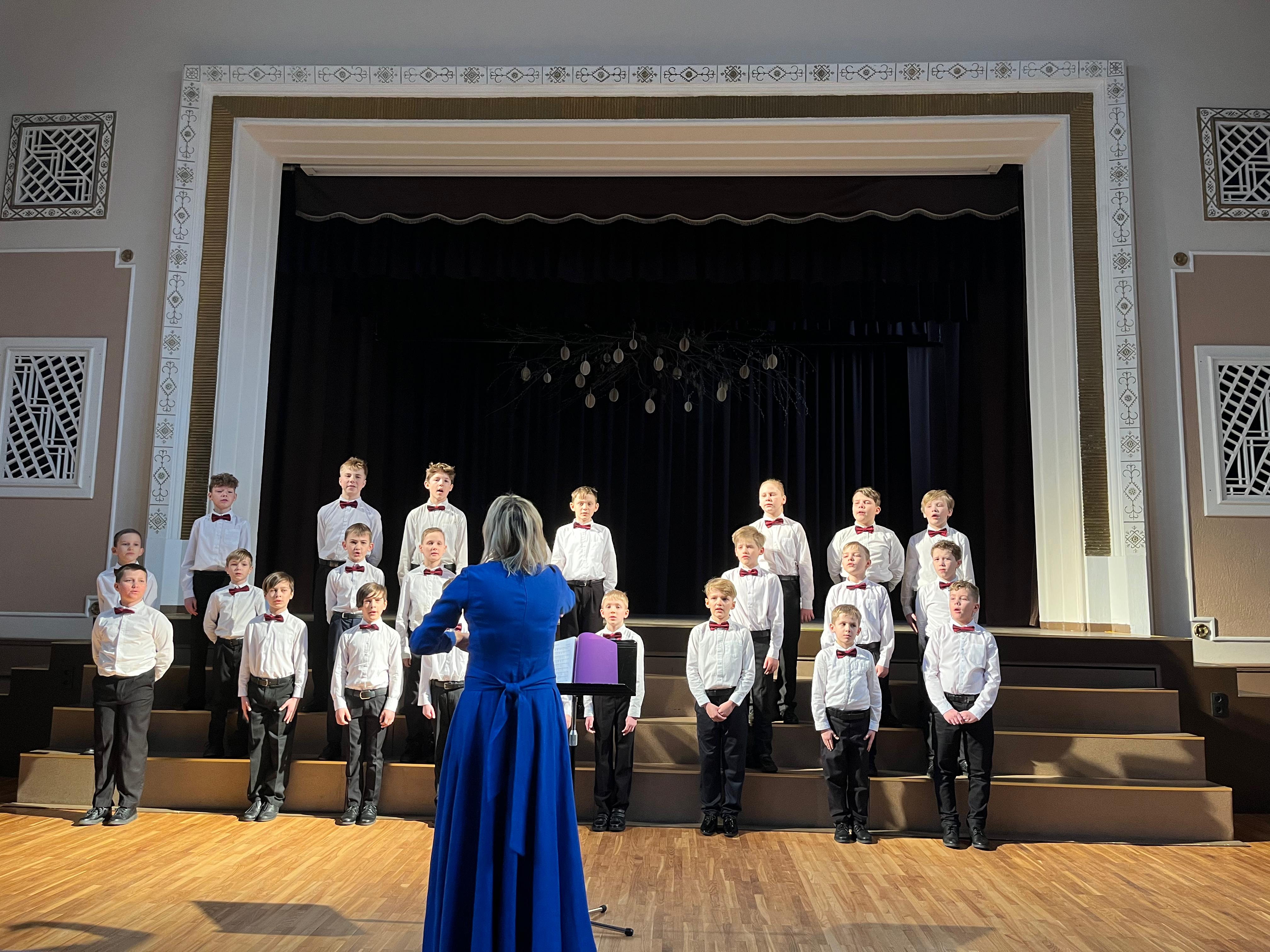 Zēnu koris baltos kreklos stāv uz skatuves, diriģē vadītāja zilā kleitā