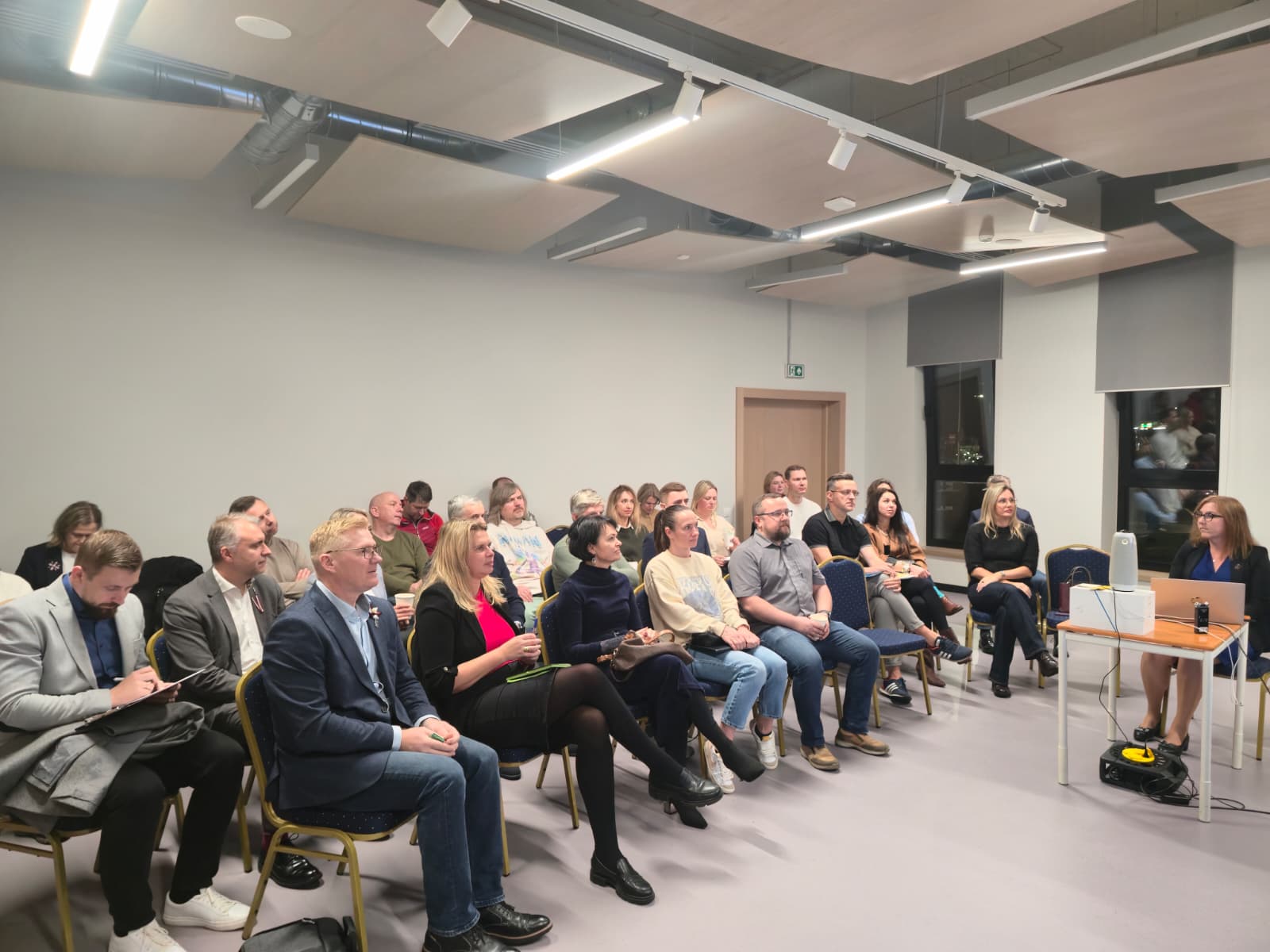 Cilvēku grupa sanāksmē sēž uz krēsliem gaišā telpā