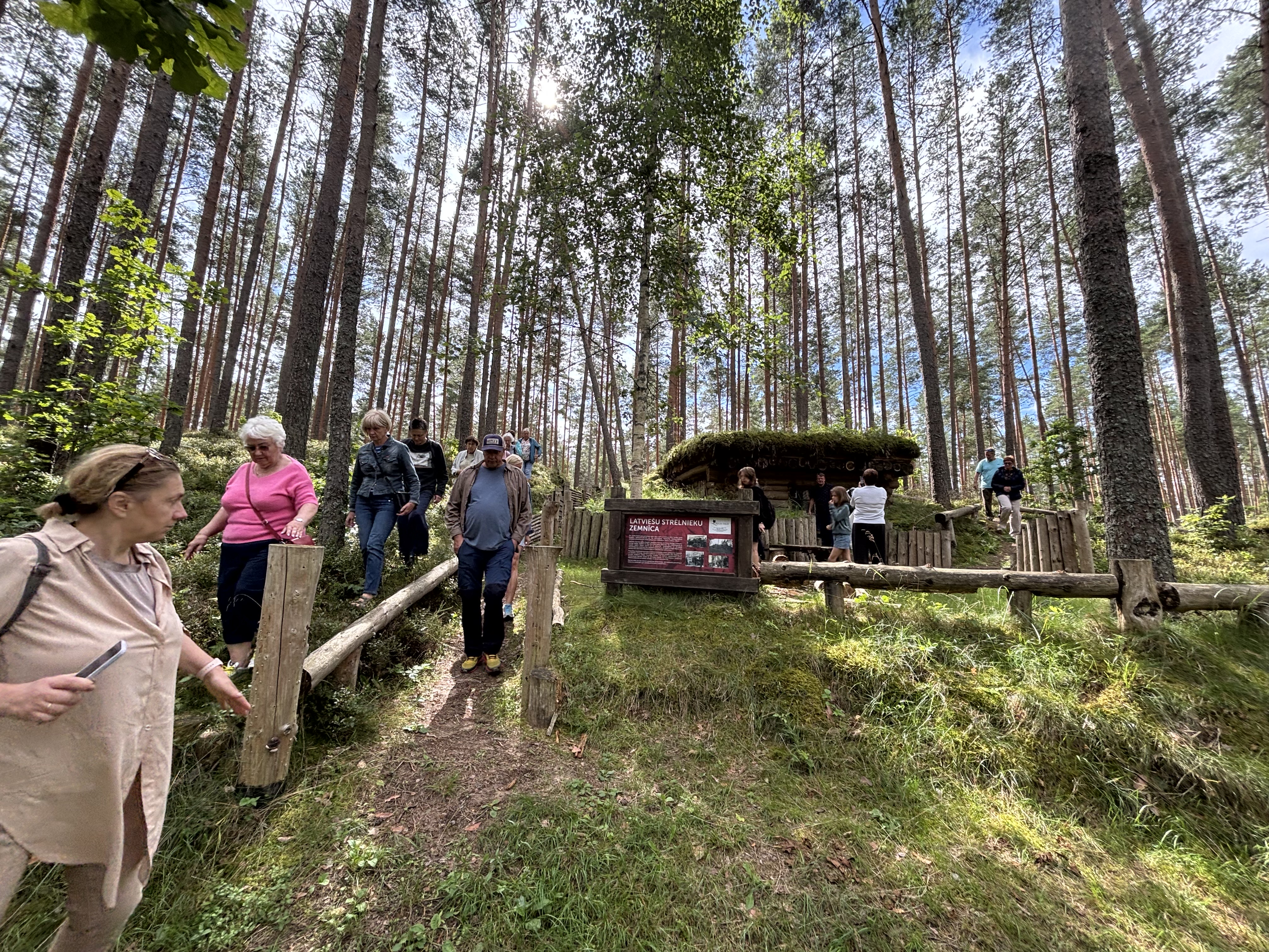 Ekskursijas dalībnieki brīvdabā apskata koka ēkas