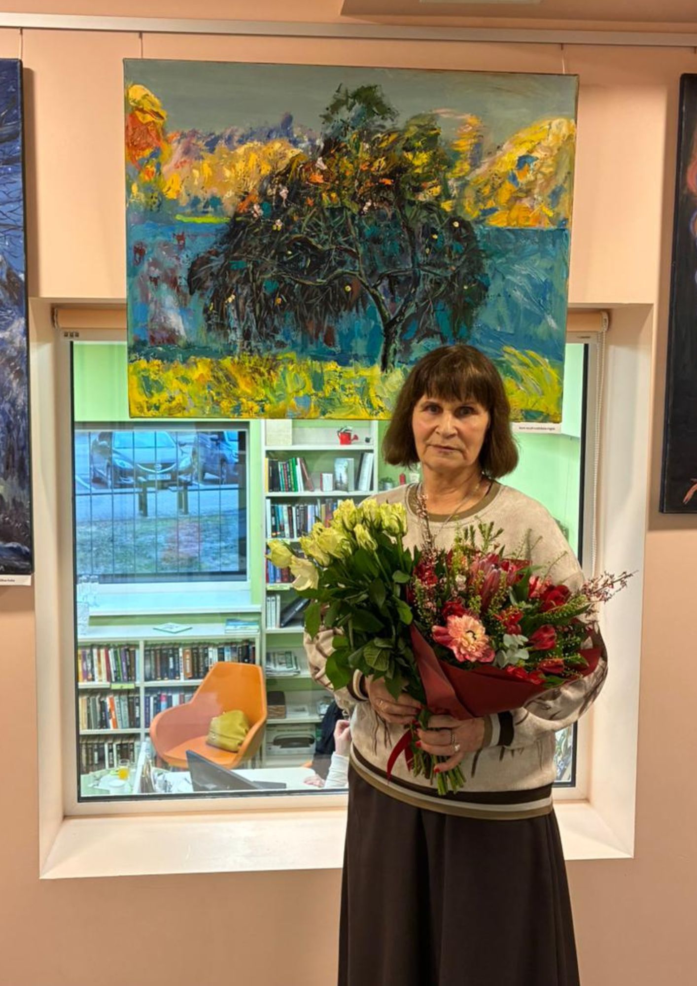 gleznotāja ar ziediem rokās pie savām gleznām