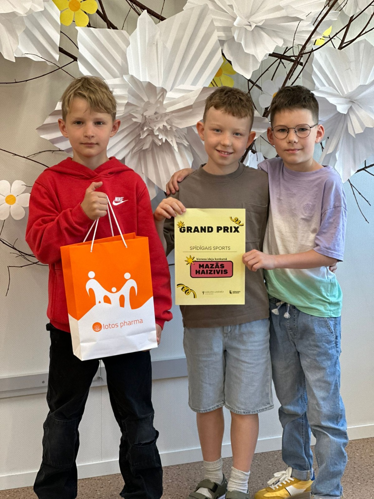 Trīs skolēni stāv pie sienas ar diplomiem rokās