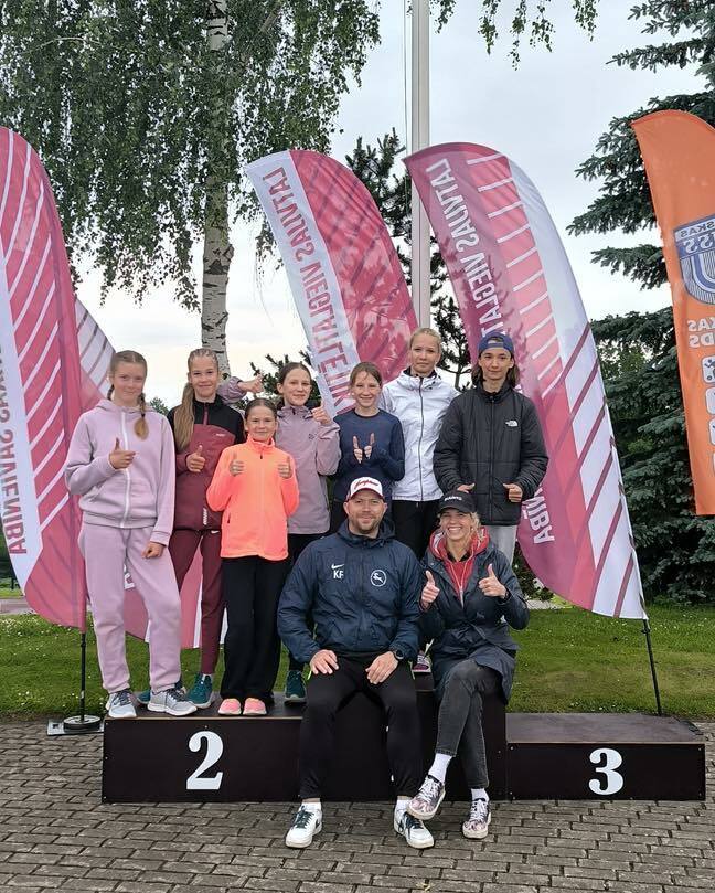 Vairāki jaunieši un pieaugušie sporta tērpos stāv un sēž uz melna pjedestāla