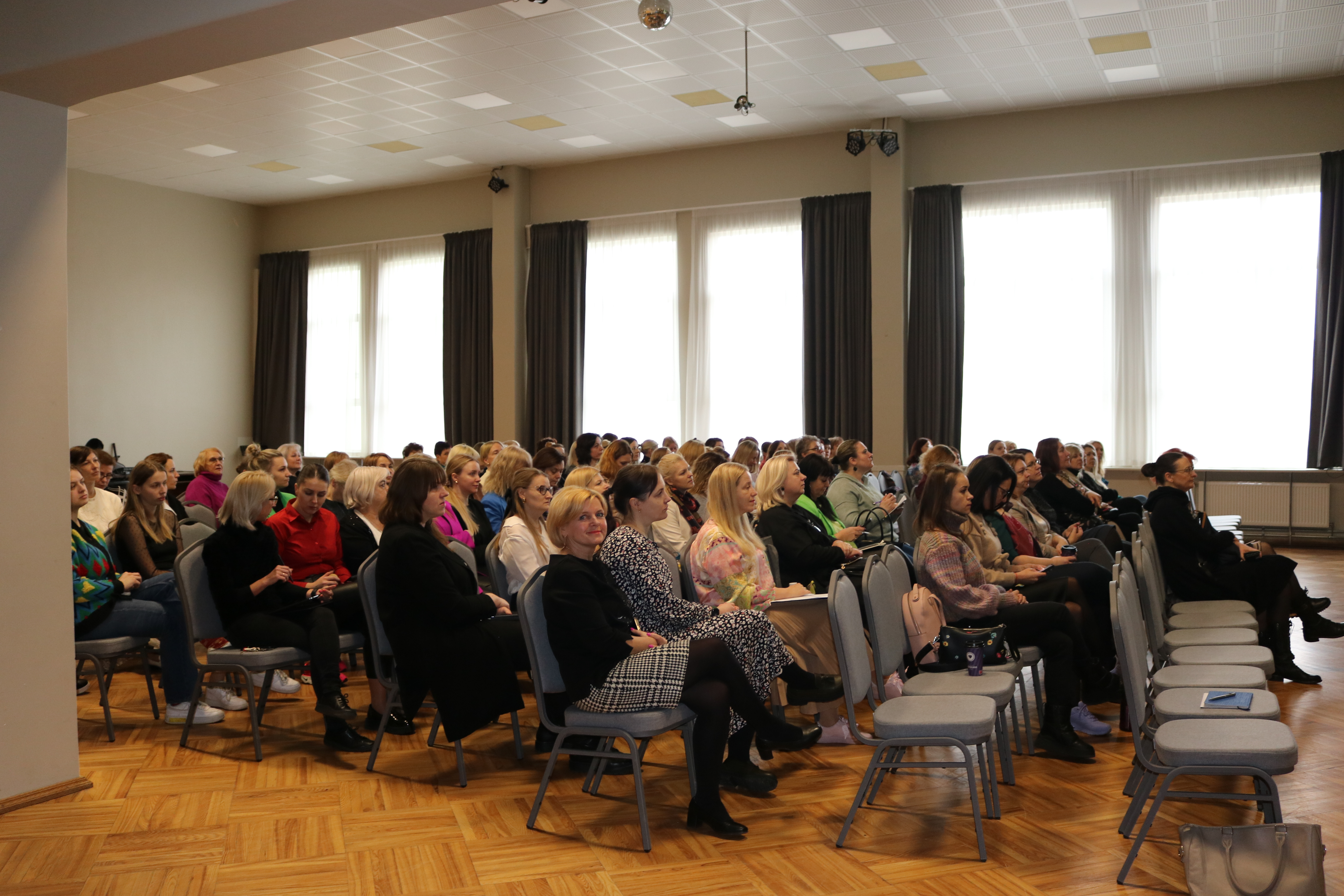 Skats uz zāli, kur krēslos sēž daudz cilvēku, klausoties konferenci