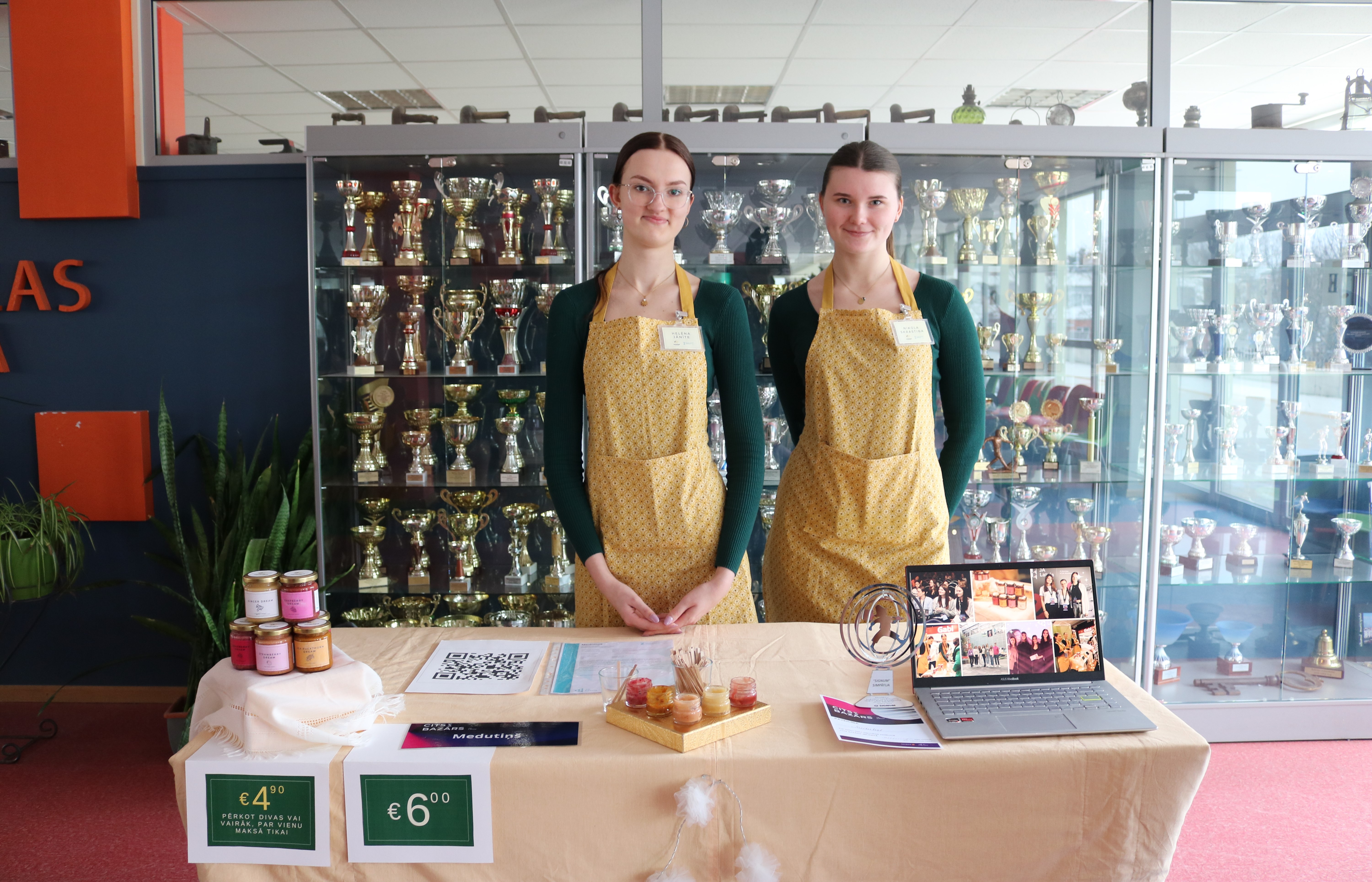 Divas skolnieces stāv pie galda, piedāvā pārdošanai savu ražoto medus produkciju