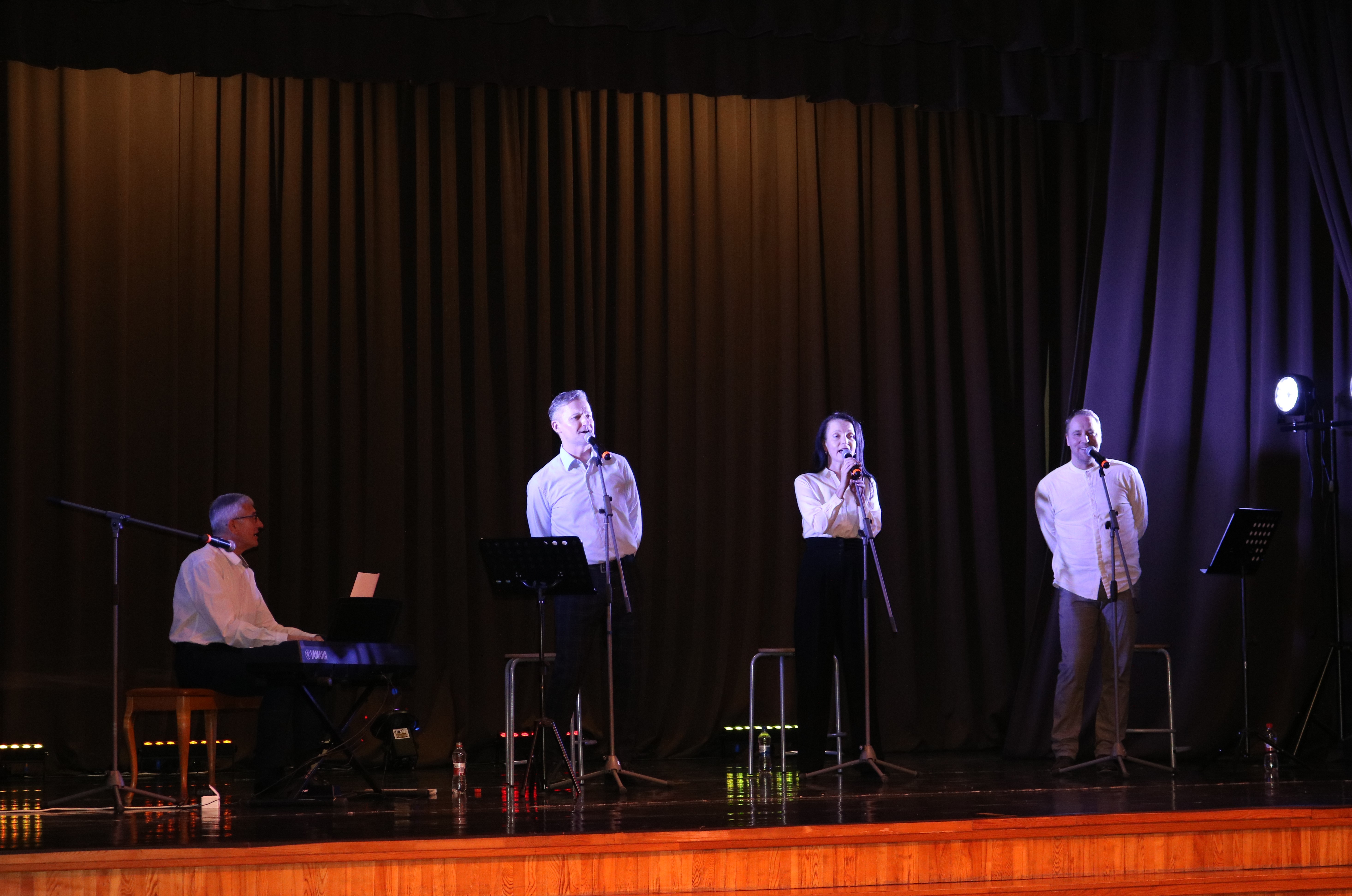 Trīs vīrieši un sieviete uz tumšas skatuves dzied