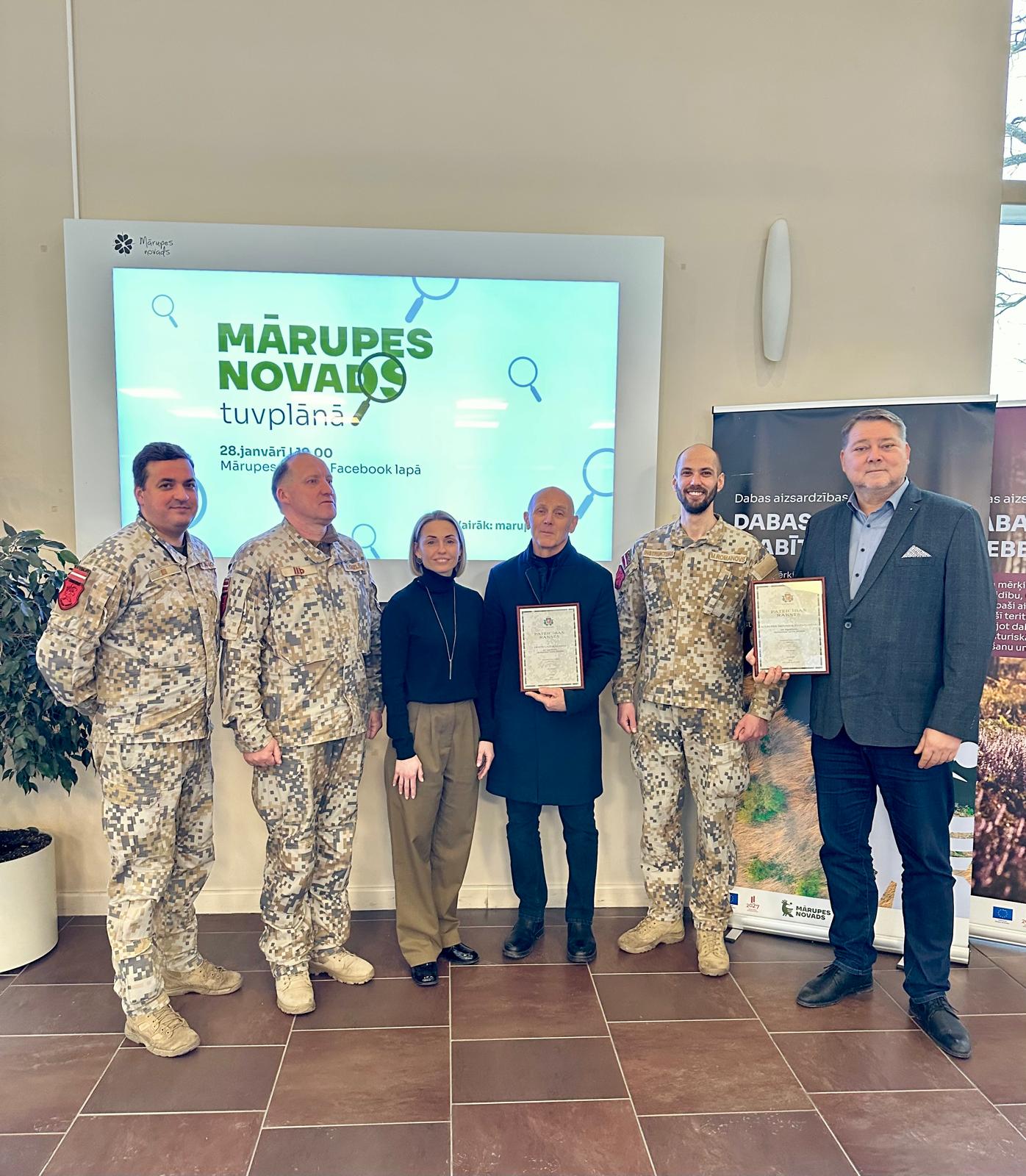 Seši cilvēki stāv kājās, aiz muguras ekrāns.