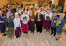 Pasākuma dalībnieku kopbilde zālē, daudz dāmas ar ziediem rokās