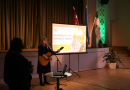 Sieviete ar ģitāru rokās dzied uz skatuves, blakus projektora ekrāns
