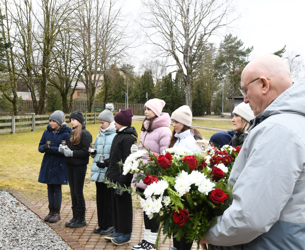 Bērni un vīrietis ar ziediem pie piemiņas akmens