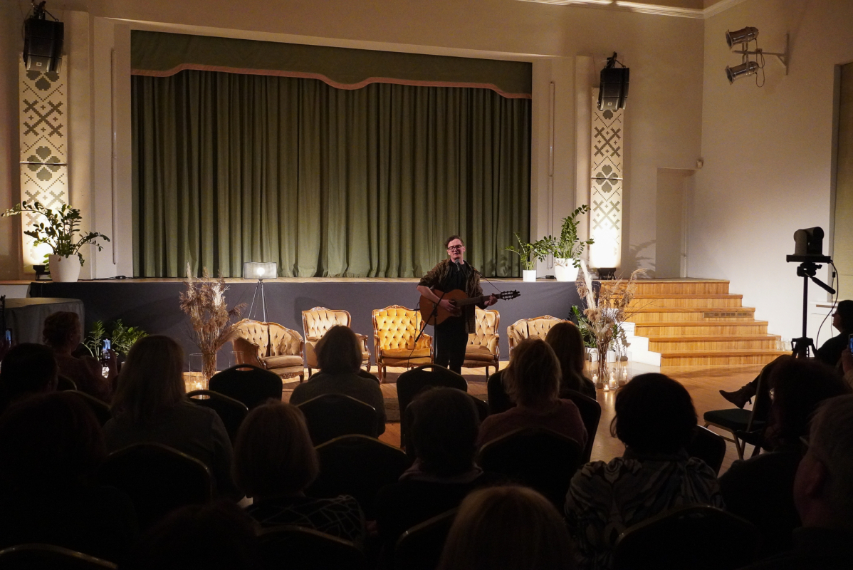 Vīrietis ar ģitāru uz skatuves un zālē sēdošie klausītāji