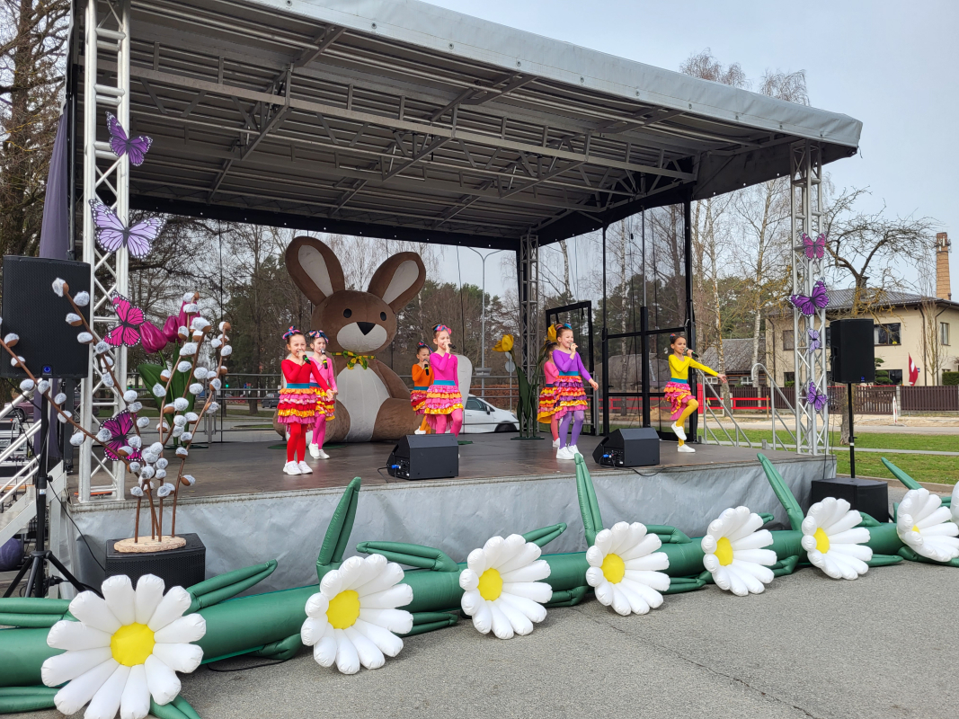 Bērnu popgrupas dalībnieki dzied uz skatuves, kas rotāta ziediem