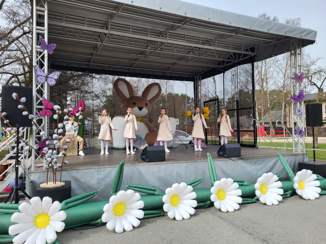 Bērnu popgrupas dalībnieki dzied uz skatuves, kas rotāta ziediem