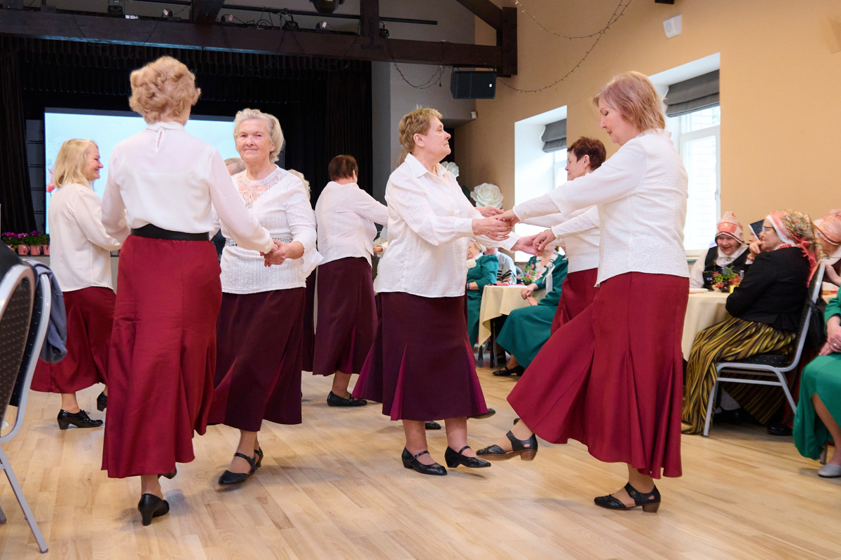 Senioru dejotājas zālē, gaišās blūzēs un sarkanos svārkos