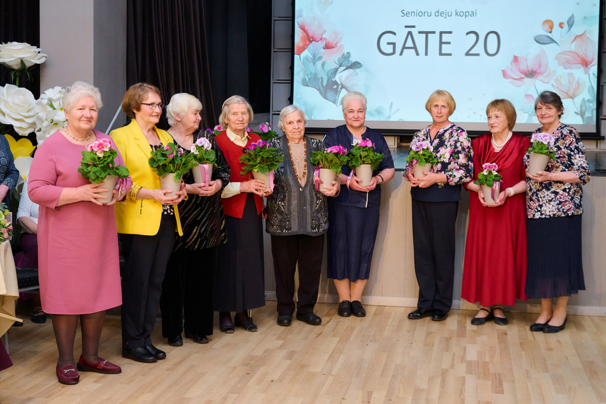 Senioru kundzes krāsainos tērpos stāv pie skatuves ar puķu podiņiem rokās