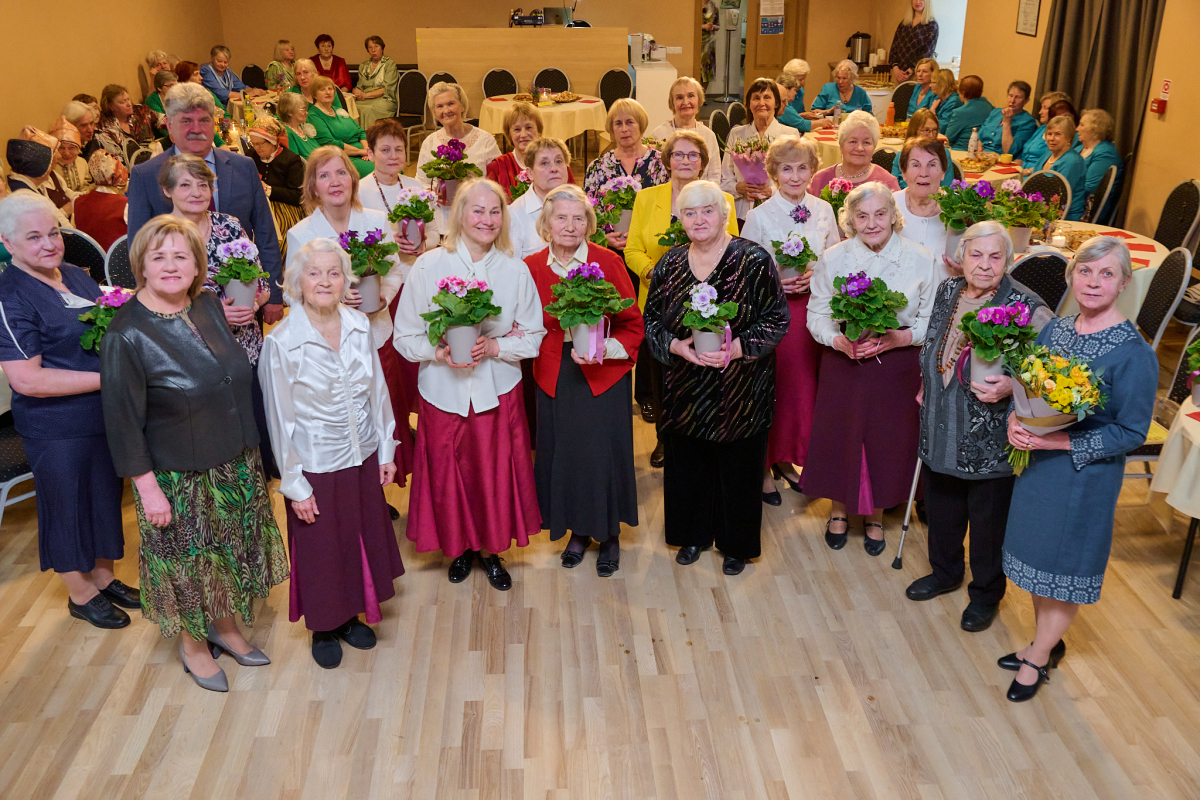 Pasākuma dalībnieku kopbilde zālē, daudz dāmas ar ziediem rokās