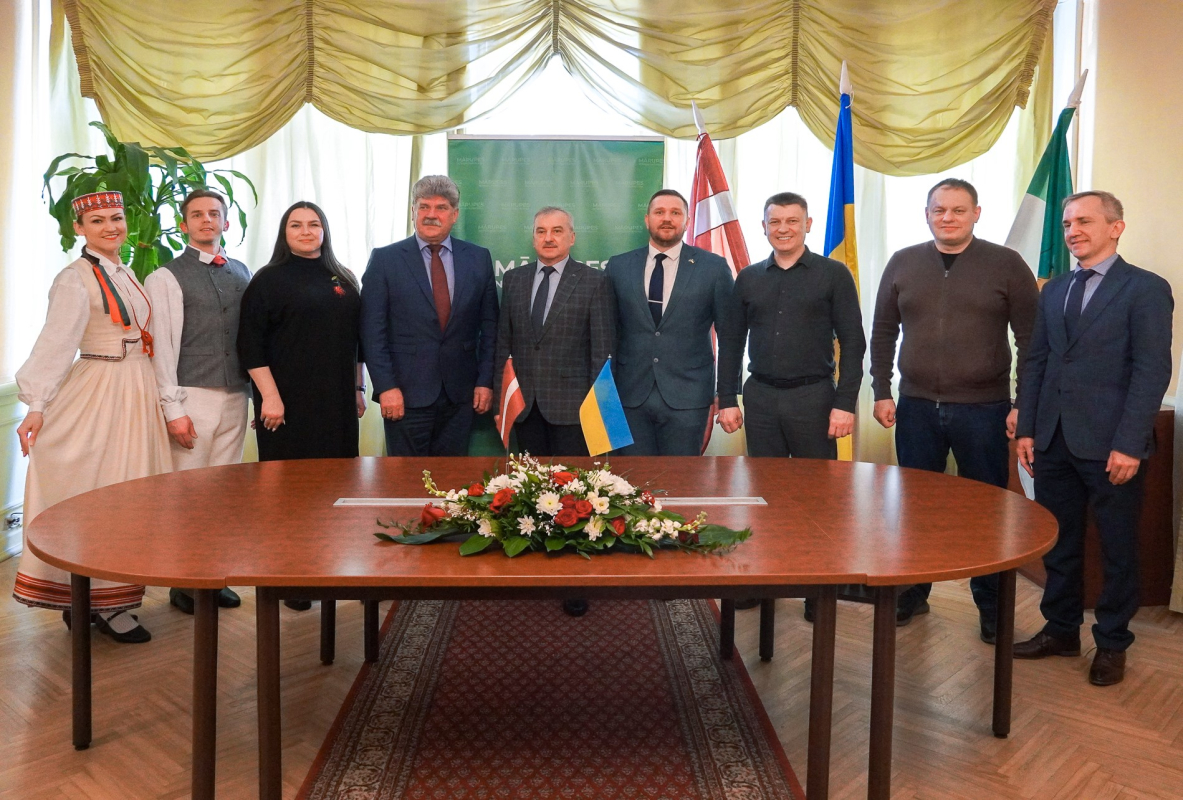 Delegācijas partneru kopbilde uz karogu fona, blakus jaunieši tautas tērpos, uz galda ziedi