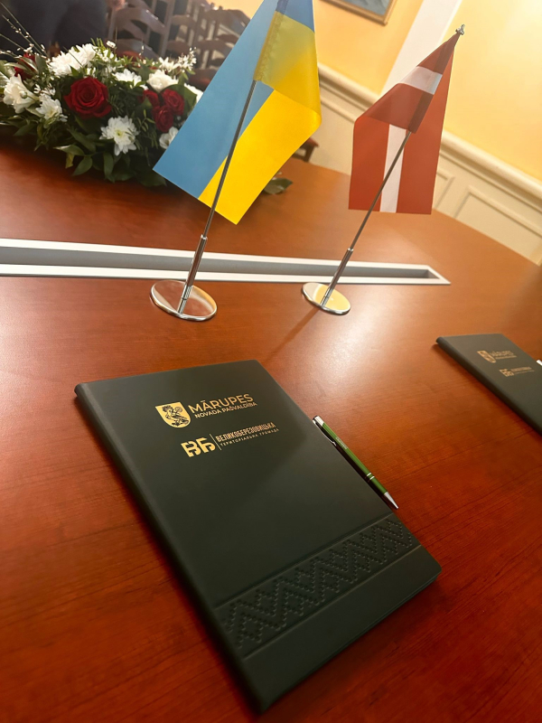 Uz galda zaļa mape ar zelta drukas ģerboņiem un divu valstu karodziņi, ziedi