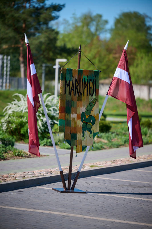 Latvijas un deju kolektīva karogi statīvā uz koku fona