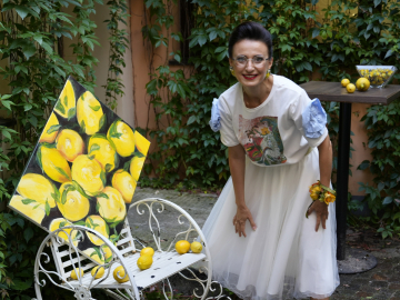Sieviete gaišās drēbēs stāv pie zaļu augu sienas, lielas gleznas ar košiem citroniem un balta soliņa, uz kura stāv citroni
