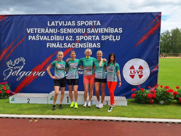 Vairākas sievietes zaļos sporta kreklos pie balta pjedestāla