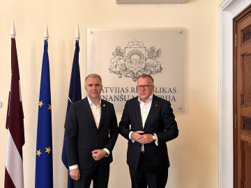 Divi vīrieši melnos uzvalkos stāv telpā pie valstu karogiem