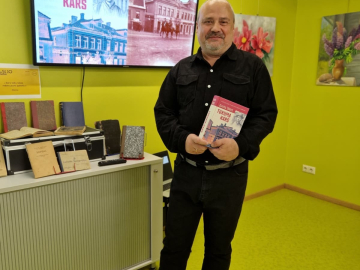 Vīrietis tumšās drēbēs ar grāmatu rokās pie dzeltenas telpas sienām