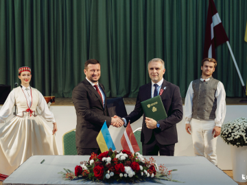 Vīrieši oficiālā darba vizītē paspiež rokas, fonā dejotāji tērpos