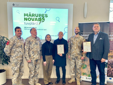 Seši cilvēki stāv kājās, fonā ekrāns