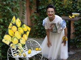 Sieviete gaišās drēbēs stāv pie zaļu augu sienas, lielas gleznas ar košiem citroniem un balta soliņa, uz kura stāv citroni