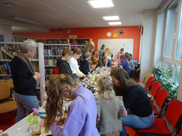 Cilvēki pulcējušies bibliotēkas telpā ap galdu, lai veidotu pavasara dekorus