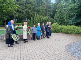 Neliels pulks cilvēku stāv ar ziediem rokās pie pieminekļa vasaras dienā