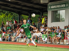 Futbols spēlētājs zaļā tērpā sit baltu bumbu futbola stadionā
