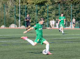 Puisis zaļā treniņtērpā sit futbola bumbu zaļā laukumā